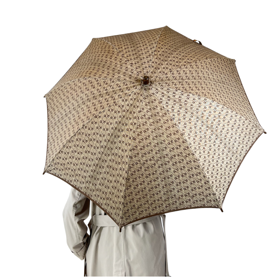 Ombrello in tessuto beige con logo gucci, manico in legno con staffa oro, tracolla staccabile.